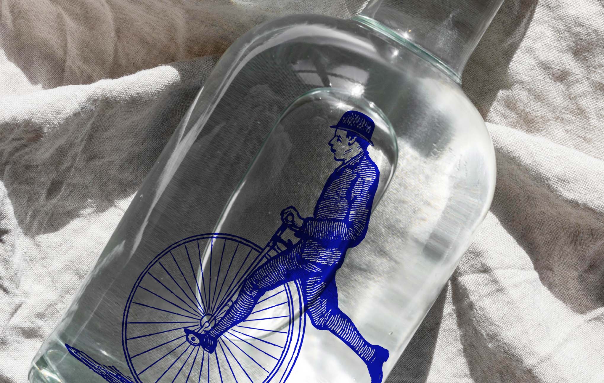 A glass bottle containing a clear spirit lies on a striped cloth. Inside the bottle, a detailed blue silhouette of a man riding an old-fashioned bicycle is displayed, creating a unique and artistic visual piece