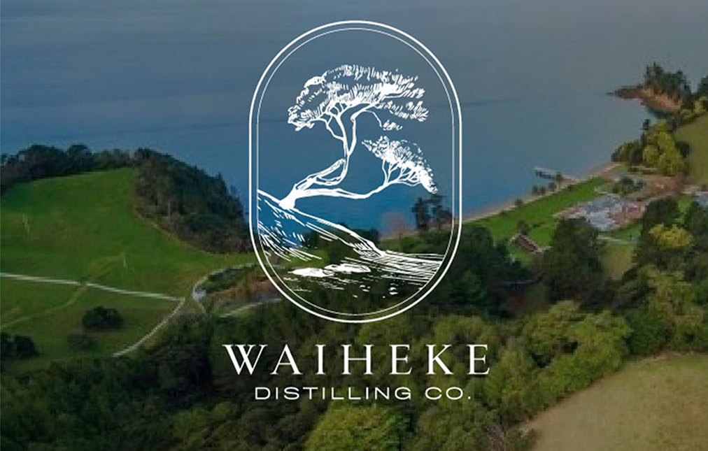 Logo of waiheke distilling co. featuring a stylized tree within a circle over an aerial view of a verdant coastal landscape with trees, fields, and a body of water. text below the logo reads 