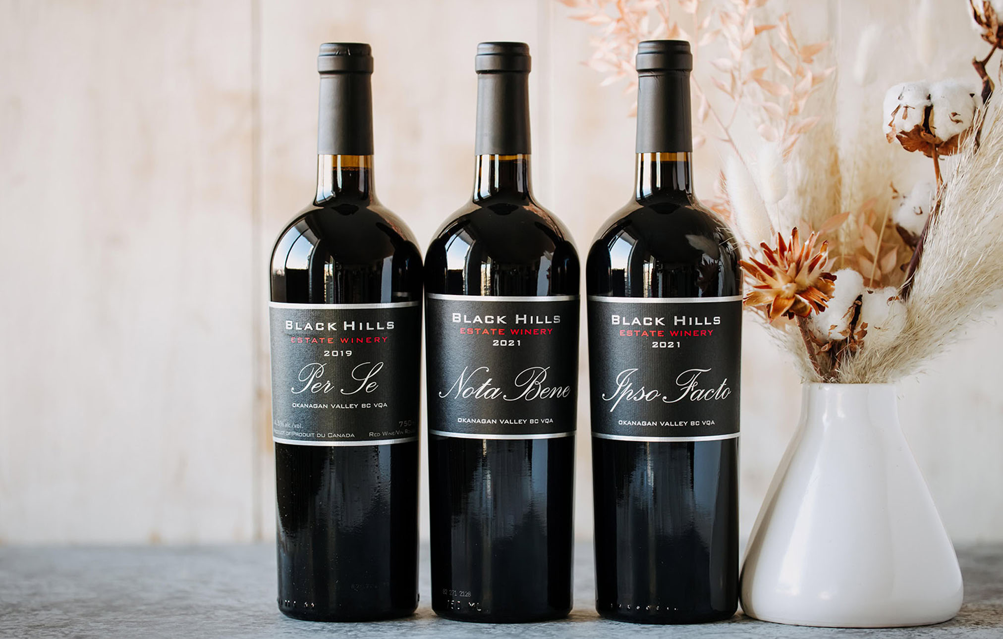 Three bottles of Black Hills Estate wine: Per Se, Nota Bene, and Ipso Facto, on a light background with a vase of dried flowers to the side.