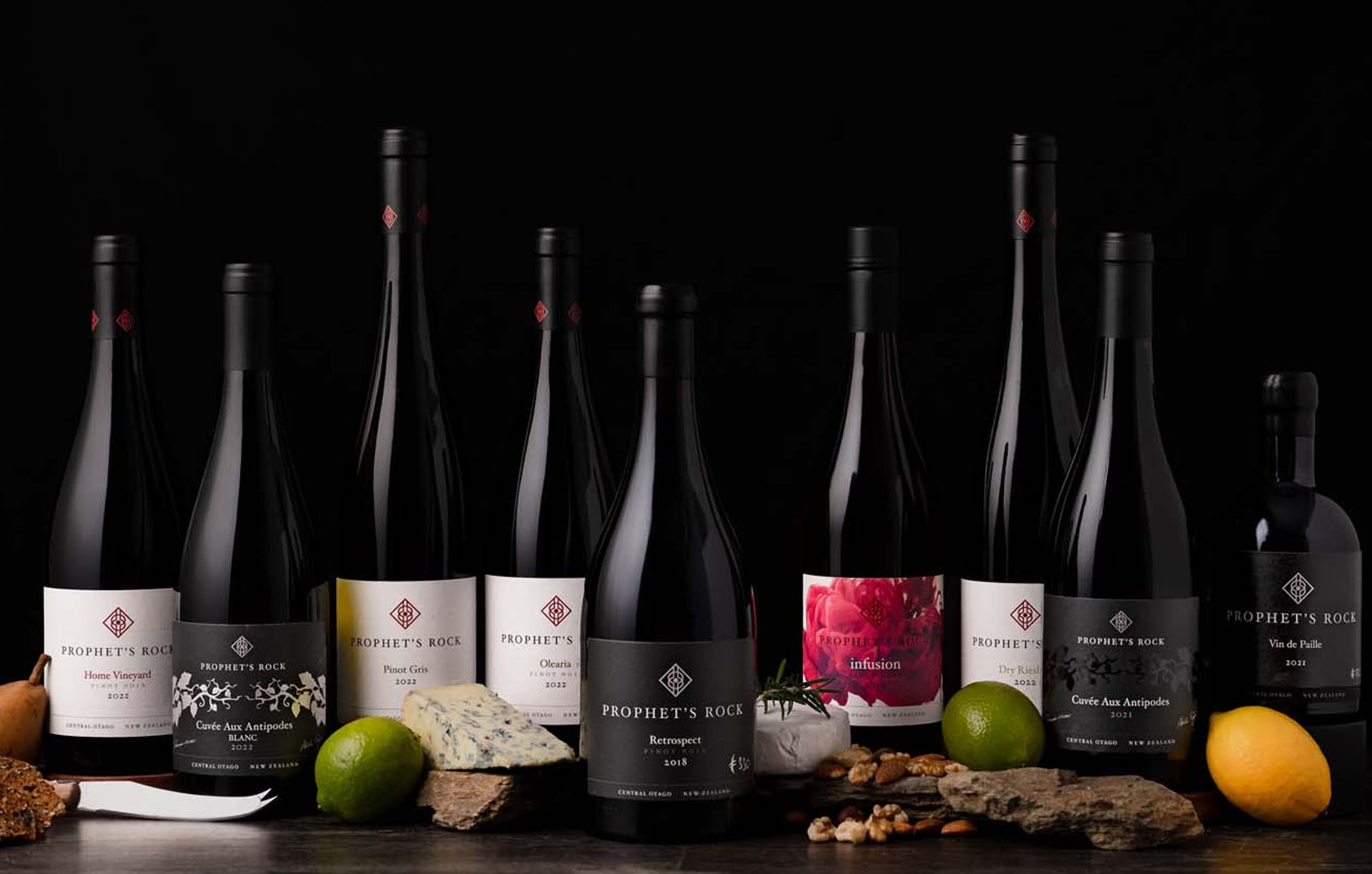 A dark and moody lineup of Prophet's Rock wine bottles, surrounded by cheese and nuts and herbs.