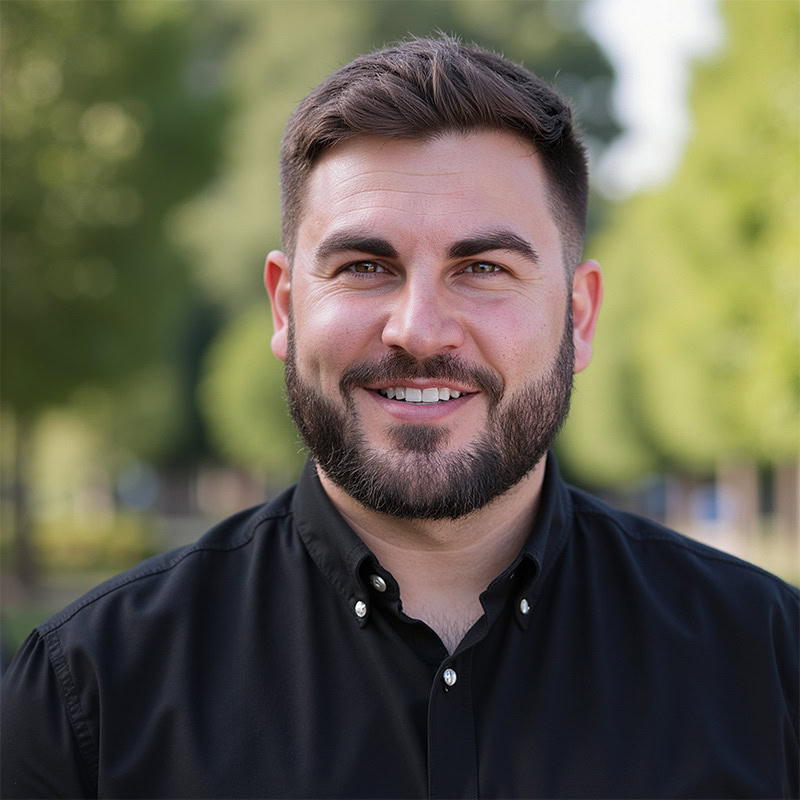 Man with dark hair and beard photographed in park setting.