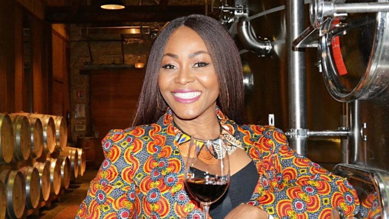 Nigerian woman with long braided hair in a winery holding a glass of wine while smiling at camera.