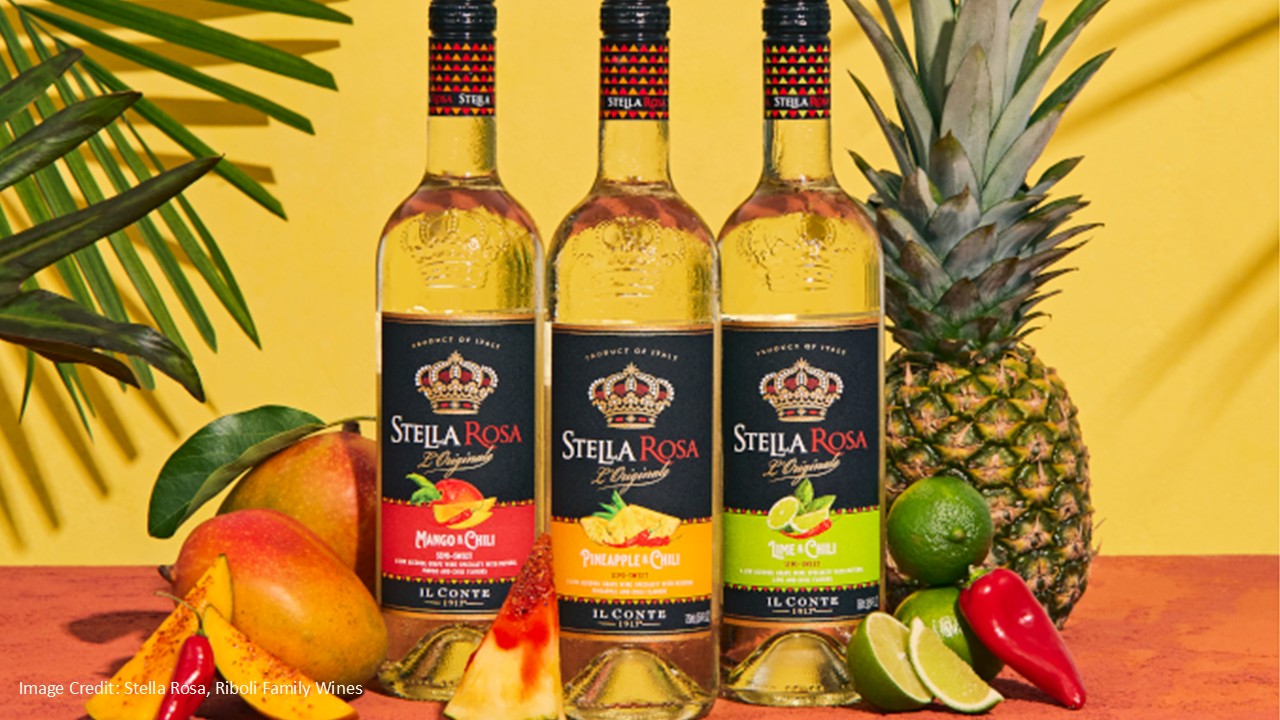 Three bottles of Stella Rosa fruit-flavored wine stand on a table surrounded by mango, pineapple, watermelon, lime, and chili peppers, with a yellow background and tropical greenery.