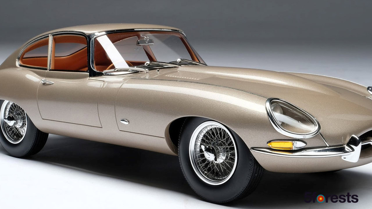 A classic beige Jaguar E-Type car with chrome detailing and wire-spoke wheels, shown in a studio setting with a gray background.