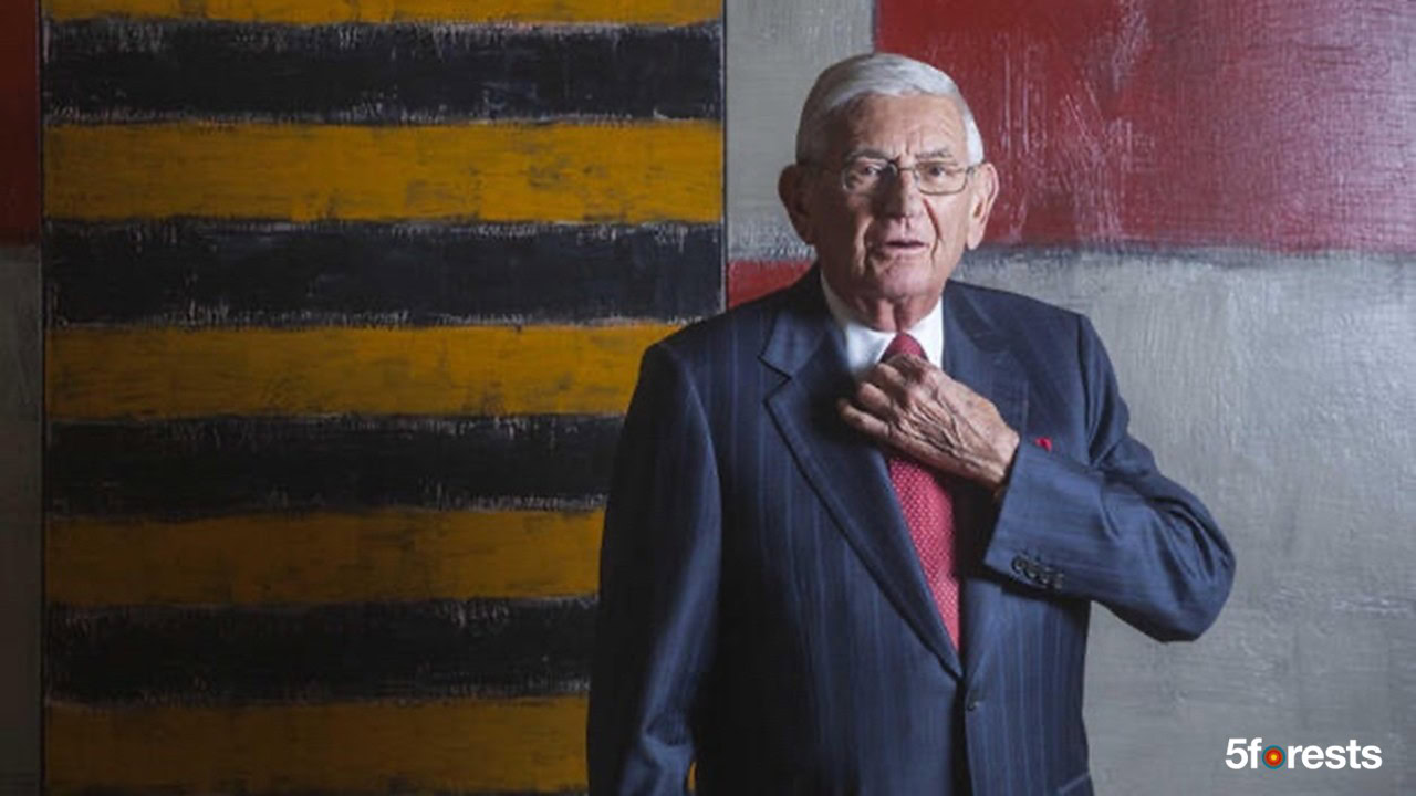 An older man in a suit and red tie adjusts his collar while standing in front of a modern, abstract painting with yellow and black stripes. 