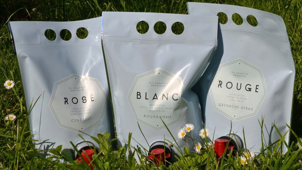 Three silver wine pouches labeled Rosé, Blanc, and Rouge stand upright on green grass with small white daisies. Each pouch features a spout at the bottom and circular handles at the top.