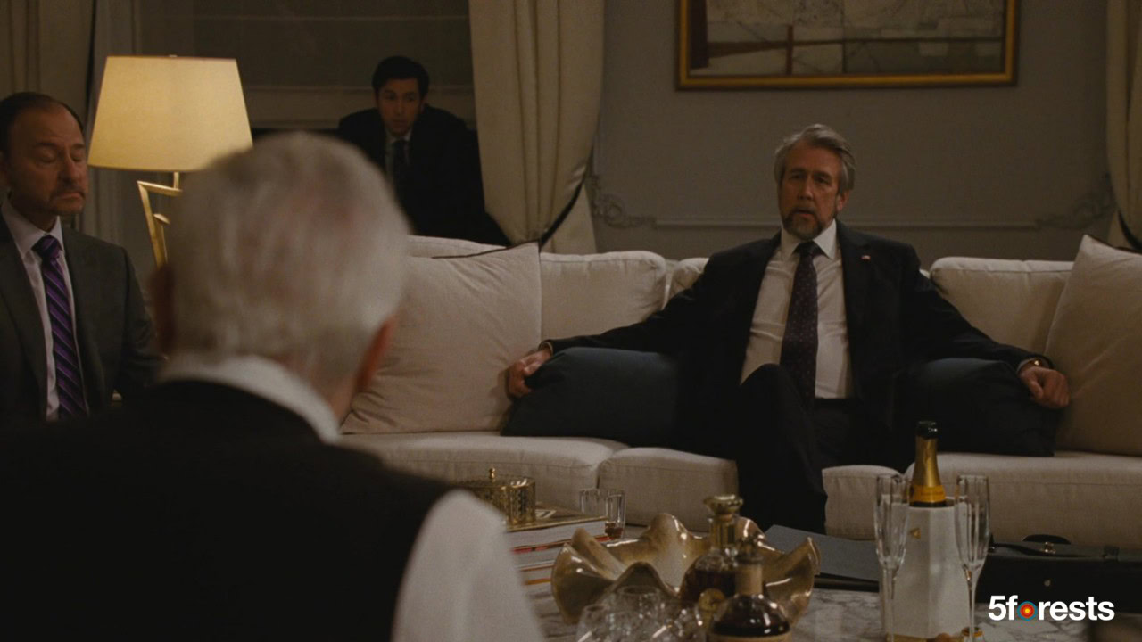 Four men in suits sit solemnly in a dimly lit, elegant living room with a white couch and coffee table displaying drinks and decorative items. One man sits apart in the background.