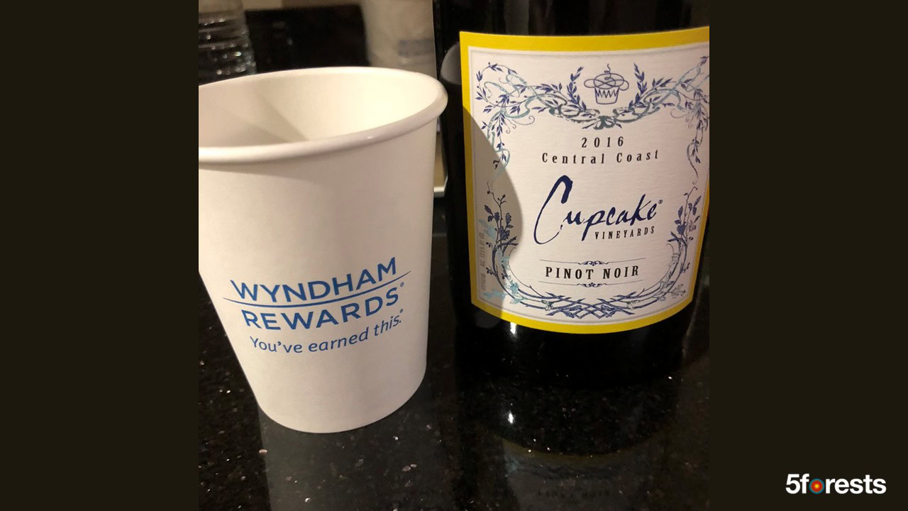A Wyndham Rewards paper cup sits next to a bottle of Cupcake Vineyards 2016 Central Coast Pinot Noir on a black countertop. 