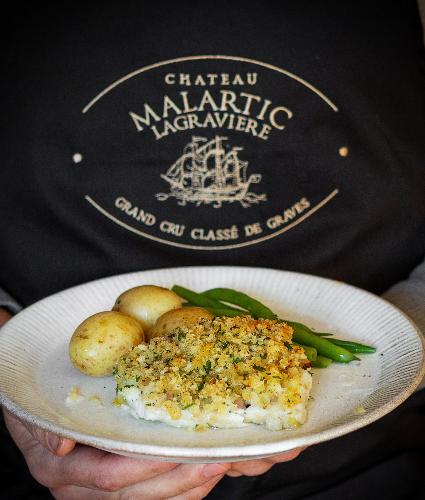A plate of fish, potatoes and green beans held in front of a Château Malartic-Lagravière apron, from The Four Seasons of Malartic
