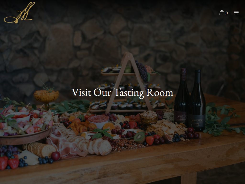 The Maysara Visit Us page in a desktop browser, headed "Visit Our Tasting Room" over a long table laid with cheese, fruit and bottles against a stone wall.