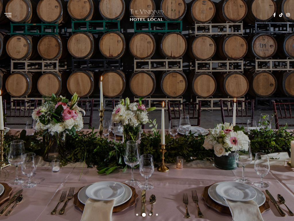 The Vineyard at Florence weddings page in a desktop browser, with a table laid with flowers, candles and glassware in front of racked barrels.